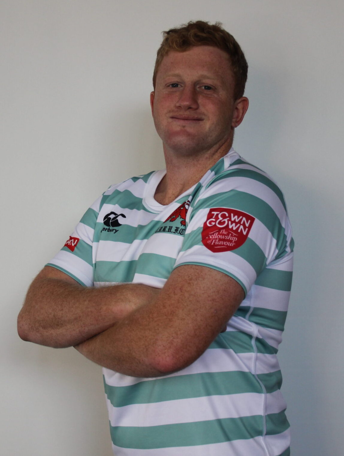 A man with short ginger hair in a light blue and white striped rugby shirt stands with arms folded, smiling slightly, in front of a plain light background—a classic look from the Cambridge rugby Varsity Matches. Logos appear on his chest and sleeve.