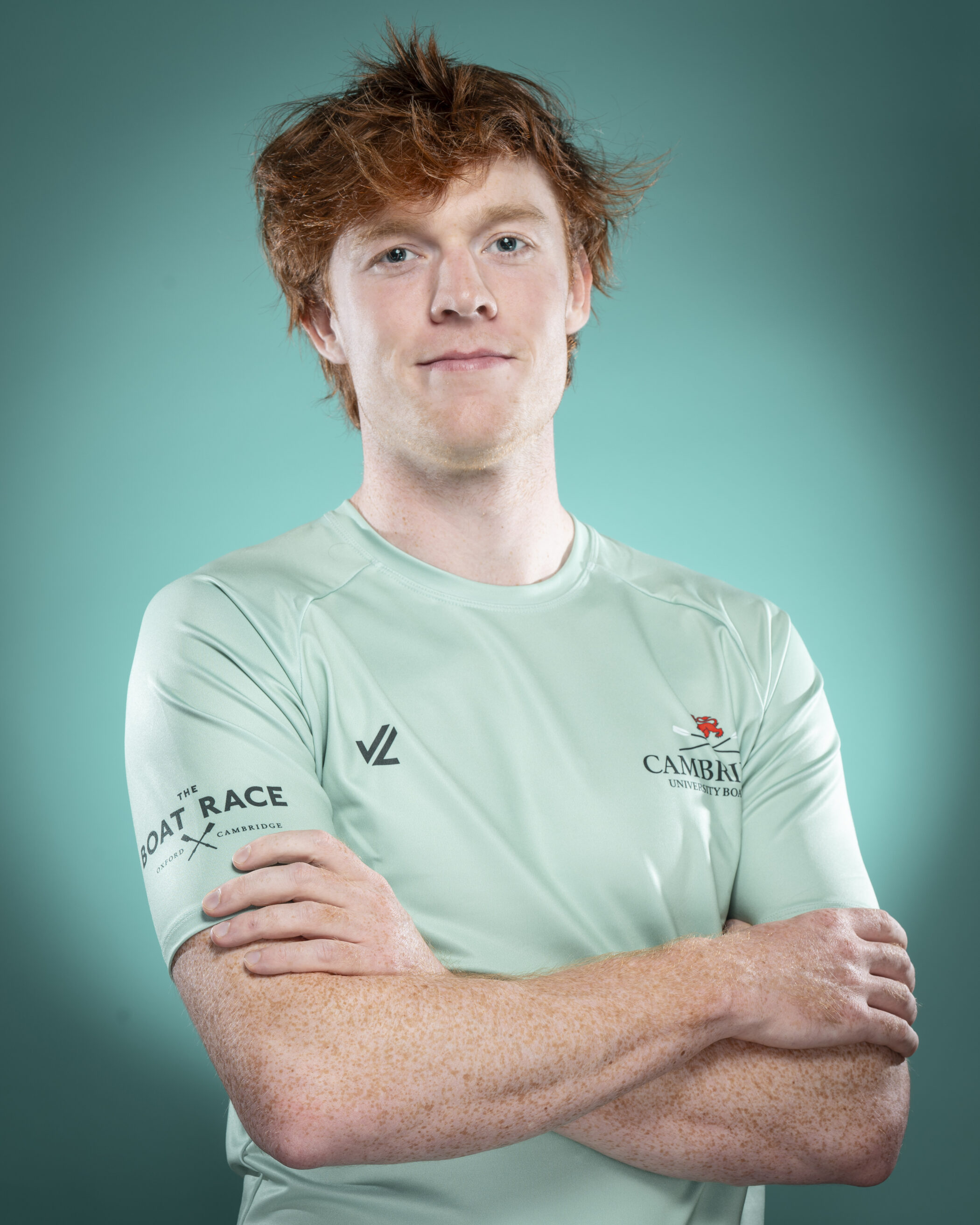 A young man with light skin and tousled red hair stands with arms folded, wearing a light green sports shirt with Cambridge University Boat Club logos, representing the spirit of college societies, against a teal background.