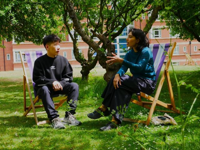 Two people sit on deckchairs under a tree, engaged in a mentoring conversation, with a brick building and green grass in the background. One gestures while speaking; both appear relaxed and attentive.