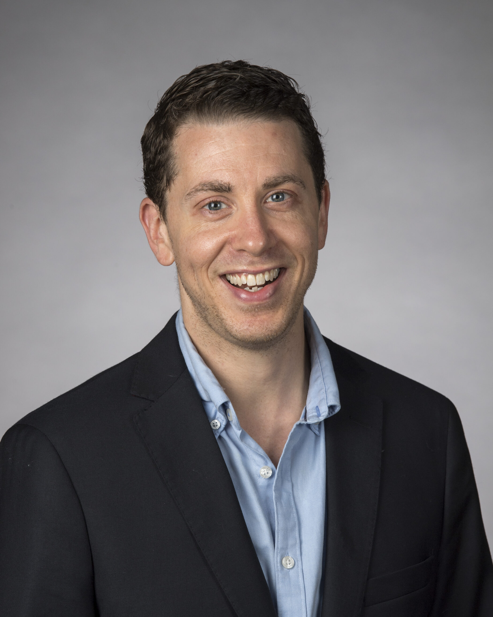 Dr Charles Leavitt, a man with short brown hair, wearing a dark blazer over a light blue button-up shirt, smiles at the camera against a plain grey background.
