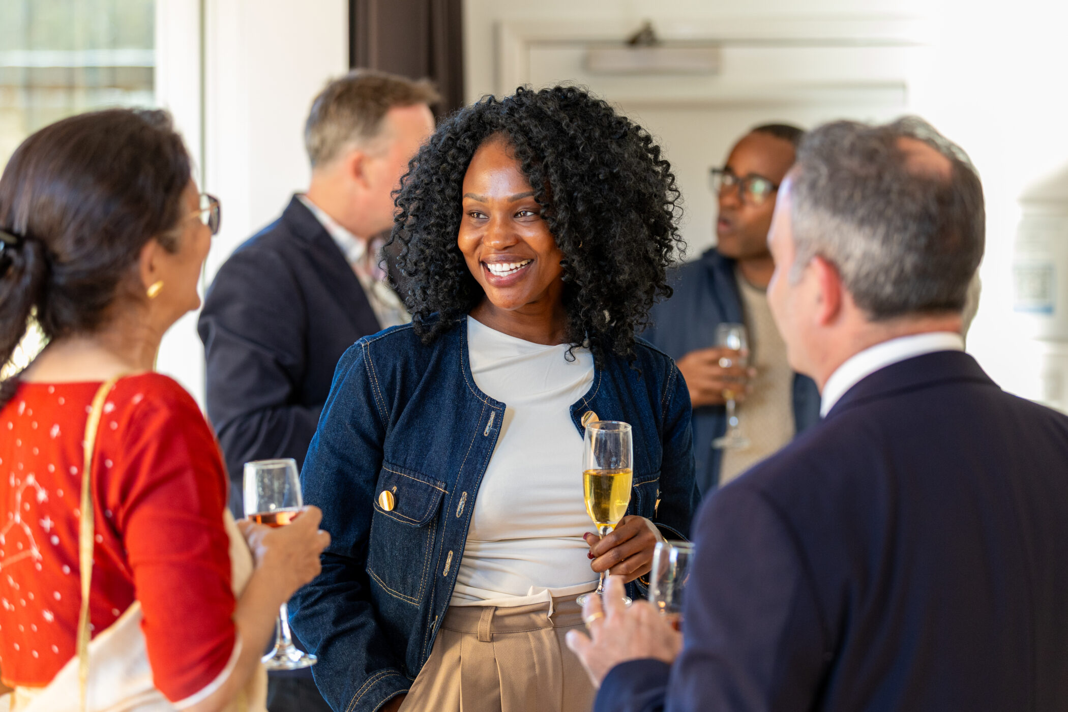 A woman with curly hair smiles whilst holding a glass of white wine at a casual indoor Von Hügel Institute event, surrounded by people in business attire socialising.