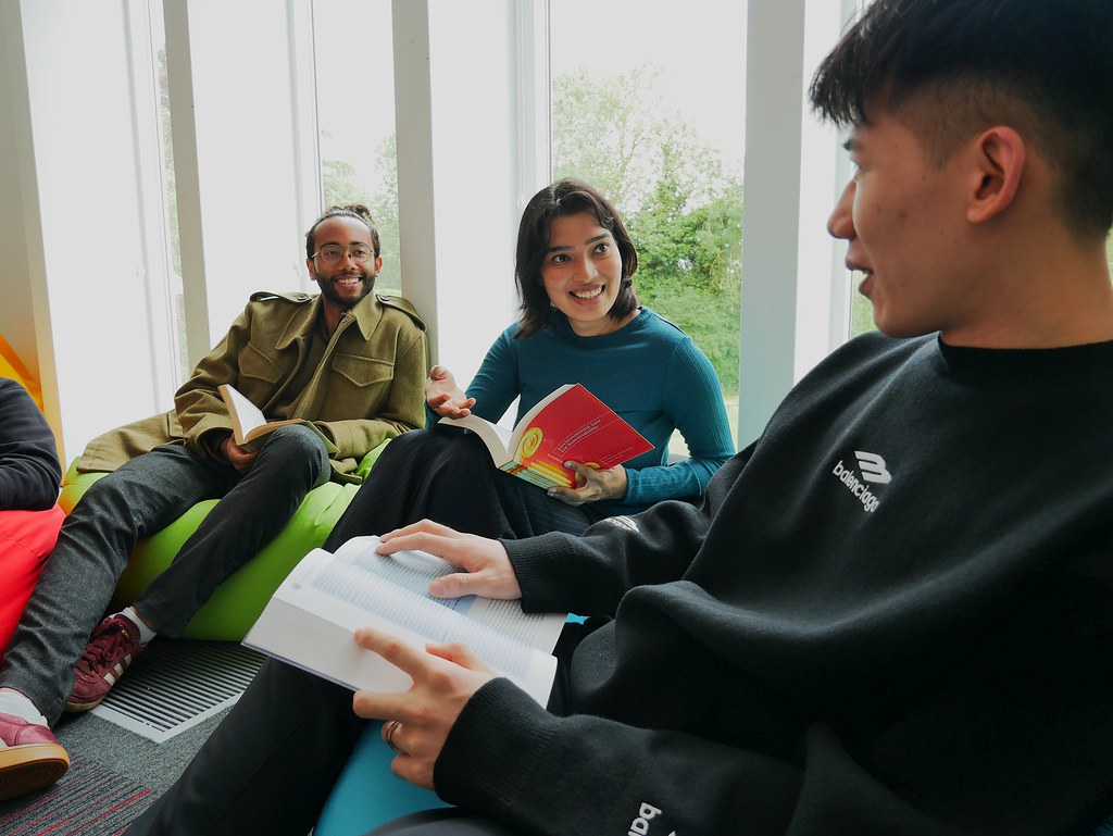 Three young adults sit together on colourful beanbags by large windows, smiling and talking whilst holding open books, suggesting a relaxed group study or discussion session focused on Linguistics and Modern Languages.